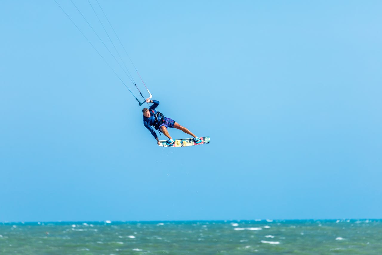 get-in-touch Kitesurfer performing an aerial trick against a clear blue sky over the ocean waves.